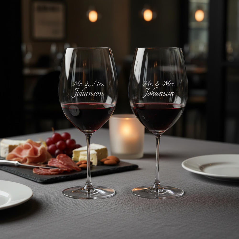 Two wine glasses with 'Mr. & Mrs. Johanson' engraving on a table with appetizers and a candle.