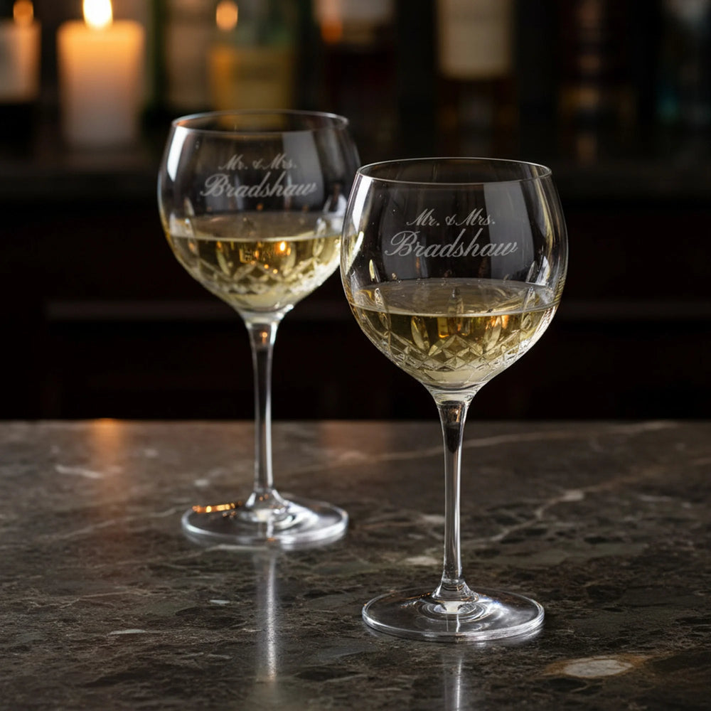 Two wine glasses with 'Mr. & Mrs. Bradshaw' engraving on a dark surface with blurred candles in the background.