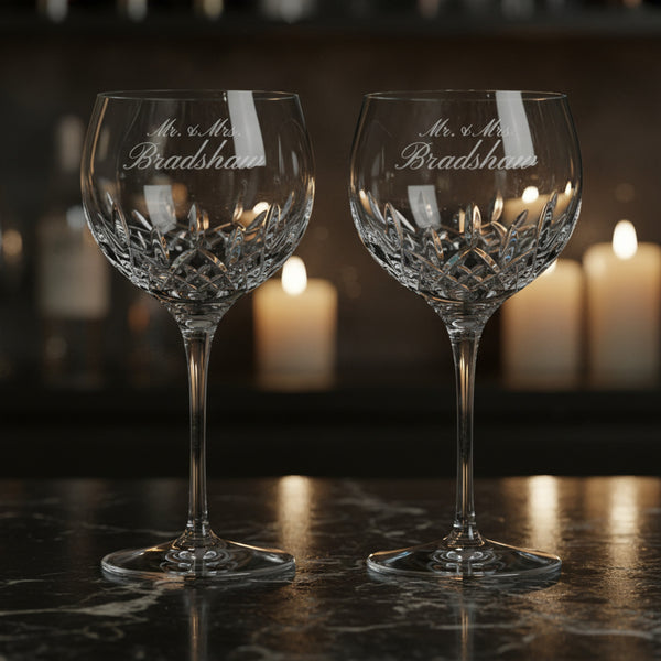 Two crystal wine glasses with engraved names on a dark surface with candles in the background.